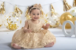 A one-year-old girl wearing a gold dress, sitting in front of her birthday cake. The cake is decorated with vibrant colors, and gold and white balloons fill the background, creating a festive atmosphere. The girl is smiling, surrounded by decorations for her special celebration.