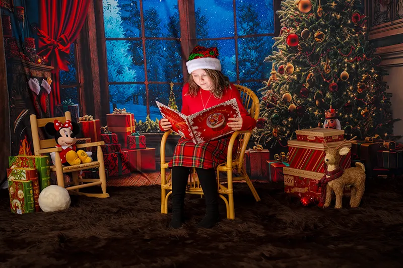 Little girl sitting on a wooden rocking chair reading a Christmas book in a cozy holiday-themed setting at Moments in Time Photography Studio