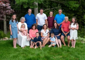 A family outdoor portrait featuring grandparents with their children and grandchildren, all smiling and posing together in a natural, scenic setting.