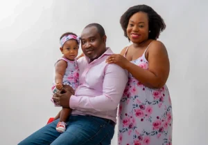 Mother and father in a Family Portrait Studio, with the father sitting on a stool holding their baby girl while the mother stands beside him, all captured in a loving and tender family moment.