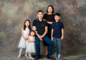 Dad sitting on a stool with his wife standing by his side and their three kids standing next to him, captured together in a family portrait at a Family Portrait Studio.