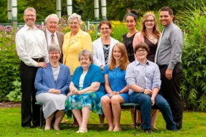 A family four-generation group photo taken at a park in downtown Halifax. The family members, ranging from a great-grandparent to young children, are smiling and standing together in a lush green park with city buildings in the background. The photo captures a moment of family togetherness and joy.