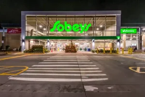 A commercial exterior photo of Sobeys Mumford Road, taken at night. The well-lit store stands out against the dark sky, with bright signage and lights illuminating the entrance. The surrounding area is visible, highlighting the store's location and vibrant presence in the evening.