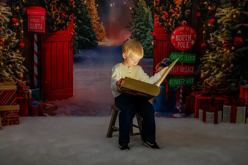 Candid photo of a young boy reading a large book in a Christmas photography backdrop with snowy ground, pine trees, and a "Welcome to the North Pole" sign.