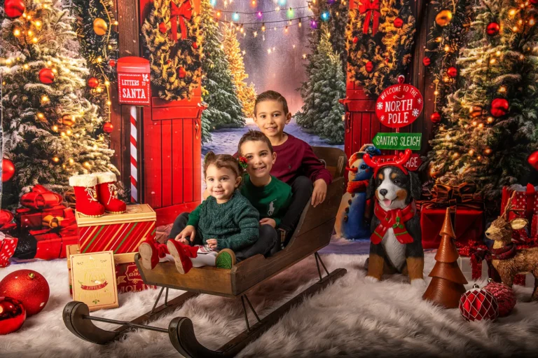 Christmas mini portrait sessions Halifax Nova Scotia 5 Three siblings in green and burgundy sitting in a wooden sleigh at a North Pole set with a Letters to Santa mailbox, Halifax