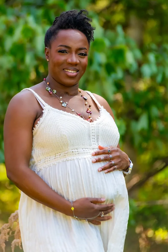 Maternity Portraits 5 Smiling pregnant woman in a cream crochet dress holding her baby bump in a sunny park, outdoor maternity portraits in Halifax