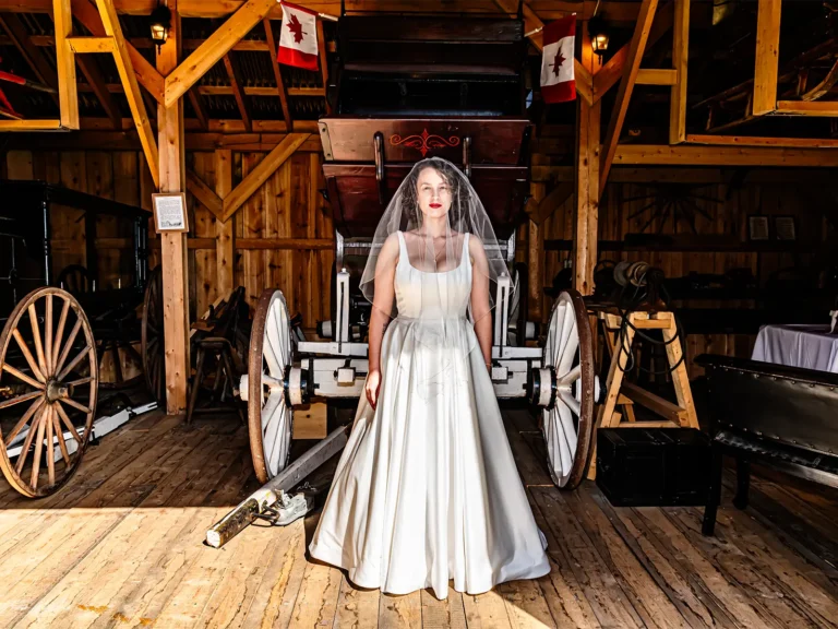 Halifax Photographers & Photography Studio 11 Bride in a white gown and veil standing in front of an antique carriage inside a rustic wooden barn for a Halifax wedding portrait