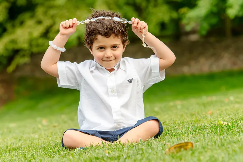 Playful outdoor portrait of a young curly-haired boy sitting cross-legged on green grass by Moments in Time Photography Studio Halifax